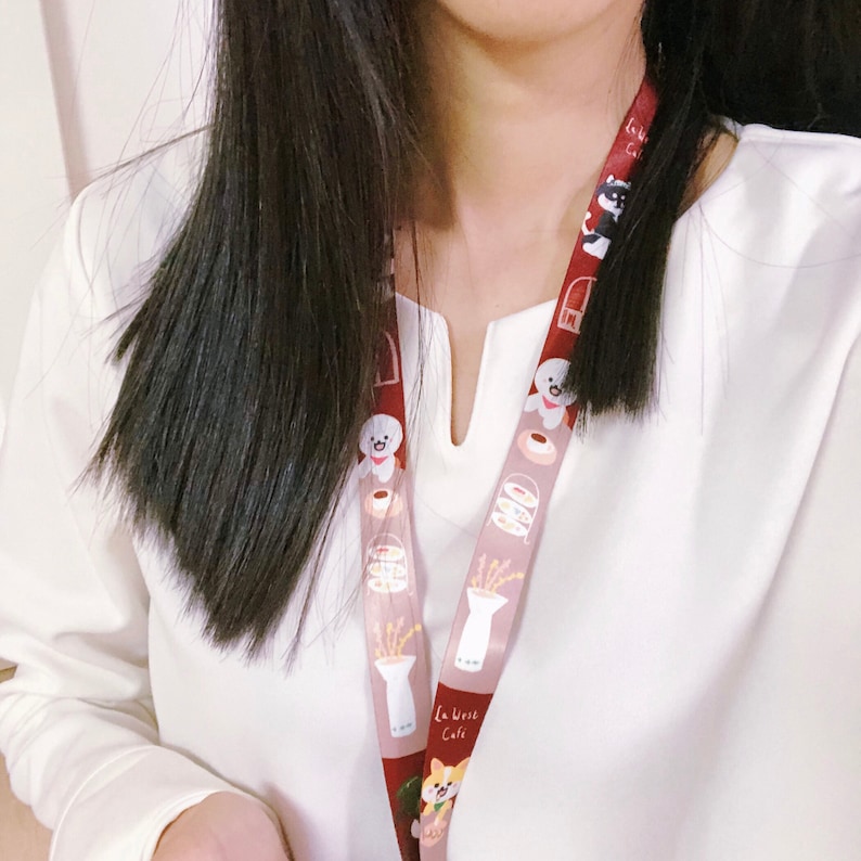 Może przedstawiać: Biała koszula z długim rękawem i dekoltem w kształcie litery V oraz brązowo-biały sznurek z kresk&oacute;wkowym psem i wzorem kawiarni. Sznurek ma nadrukowany tekst "La Vrise Cafe".