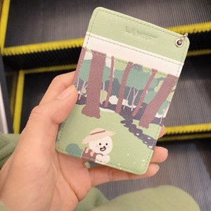 May include: A light green rectangular card holder featuring a printed forest scene with brown trees and a small white character wearing a straw hat. The accessory includes a white horizontal slot and a metal eyelet for a keychain attachment.