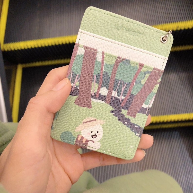 May include: A light green rectangular card holder featuring a printed forest scene with brown trees and a small white character wearing a straw hat. The accessory includes a white horizontal slot and a metal eyelet for a keychain attachment.