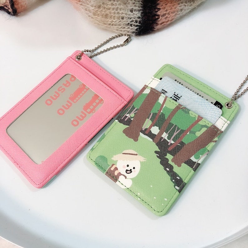 May include: Two rectangular card holders with silver ball chains. One is solid pink with a clear window displaying the text PASMO and a bus icon. The other is light green with a graphic of a cartoon character in a hat in a forest.