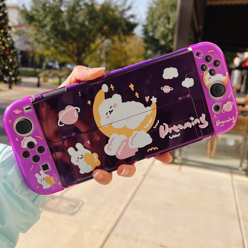 May include: Purple and white Nintendo Switch case with a cartoon design featuring a sleeping bear, a crescent moon, and stars. The case has the word "Dreaming" written on it.