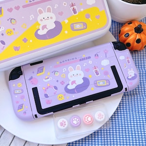 May include: A lavender Nintendo Switch case and controller with a cute bunny design. The case features a bunny on a cloud, along with other graphics. Includes matching thumb grips with paw prints. A second case is visible in the background.