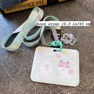Op de afbeelding: Een licht muntgroene ID-badgehouder met een bijpassend koord met een herhalend patroon van kleine dieren en vormen. De houder toont een cartoonkonijn en een kat, met een doorzichtige spiraalbevestiging en een lange riem van 43 cm.