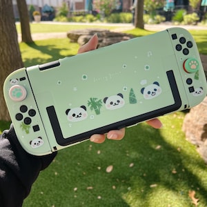 May include: A pale green Nintendo Switch case decorated with panda bear illustrations and the text "Sunny Friends". The case has black buttons and a paw print design on the joystick. The case is held in front of a green grassy background.
