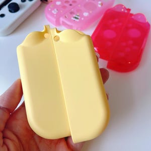 May include: A light yellow protective case for a gaming console, with a rounded top and a smooth surface. Other cases in pink and red are visible in the background.