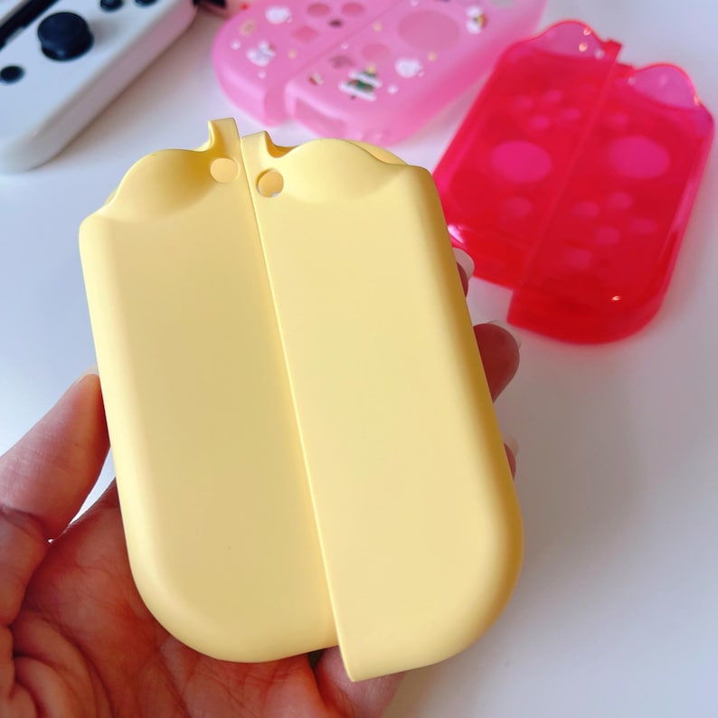 May include: A light yellow protective case for a gaming console, with a rounded top and a smooth surface. Other cases in pink and red are visible in the background.