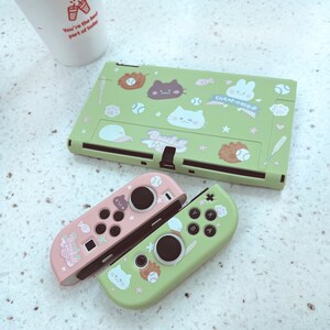 May include: A green and pink Nintendo Switch console with a baseball themed design. The console is decorated with cartoon characters and the words "Baseball Championship". The console is accompanied by two controllers, one pink and one green.