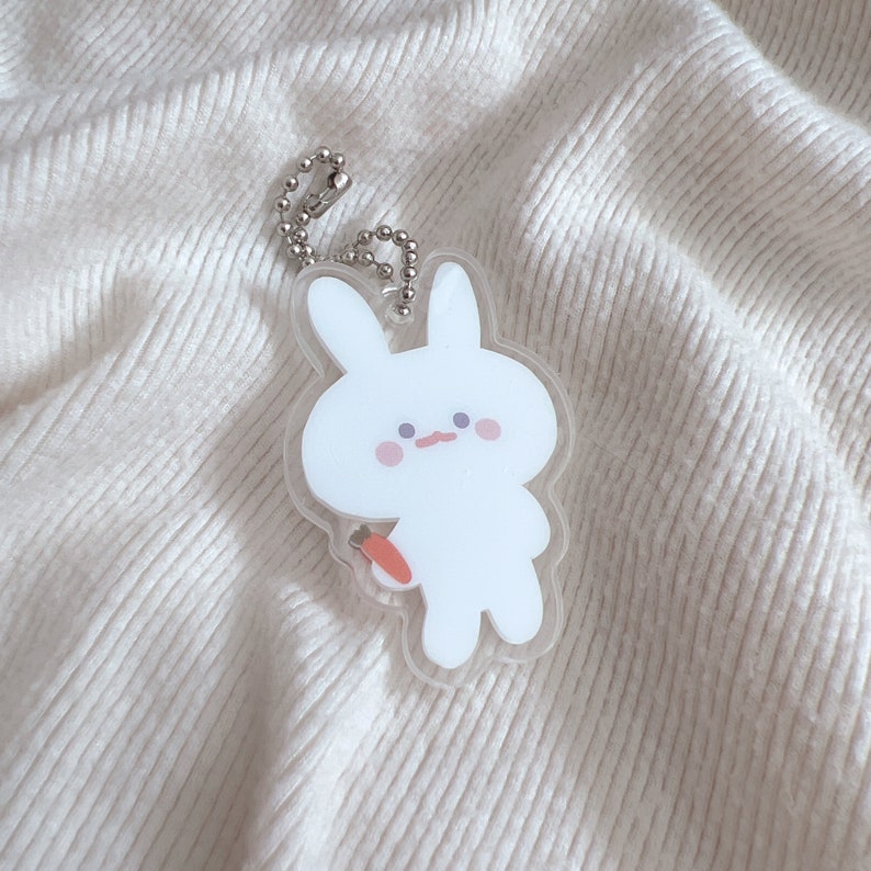 Op de afbeelding: Een witte konijnen sleutelhanger met een wortel in zijn hand. De sleutelhanger is gemaakt van helder acryl en heeft een zilveren ketting.