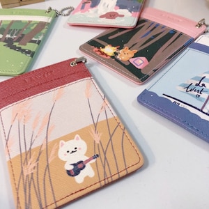 May include: A collection of five rectangular card holders featuring various graphic illustrations, including a white cat playing a guitar in a field, a campfire scene, a sailboat with the text 'la west', and forest landscapes, each with a metal attachment.