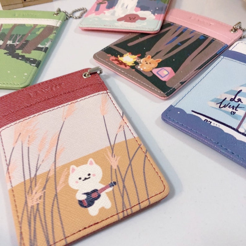 May include: A collection of five rectangular card holders featuring various graphic illustrations, including a white cat playing a guitar in a field, a campfire scene, a sailboat with the text 'la west', and forest landscapes, each with a metal attachment.