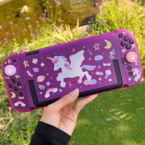 May include: A purple Switch console case featuring a white unicorn with wings, stars, and clouds. The case has pink paw print designs on the control buttons. The case is designed for a gaming console.
