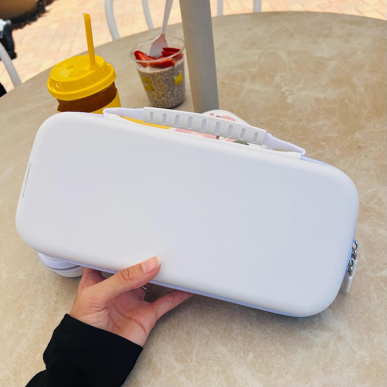 May include: A white rectangular case with a handle and a silver clasp. The case has rounded edges and a light blue trim. A yellow drink cup with a straw is visible in the background.