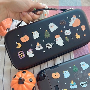 Pode incluir: Estojo de transporte preto com alça, decorado com adesivos temáticos de Halloween. Os adesivos incluem abóboras, fantasmas, árvores e a palavra "BOO" em um balão. O estojo é projetado para um console de jogos.