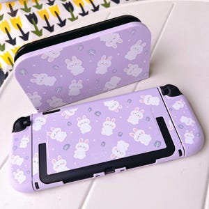 May include: A lavender Nintendo Switch console and dock set. The set features a repeating pattern of white cartoon bunnies and small green leaves. The text "Starry Tales" is printed on the console. The dock is black on top.
