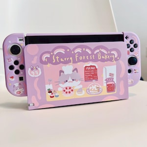 May include: A purple Nintendo Switch case with a cute cartoon cat wearing a chef's hat and a bakery sign that says "Starry Forest Bakery".