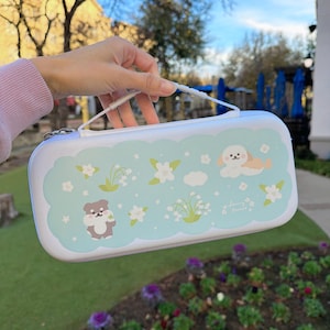 Pode incluir: Uma mala de transporte azul claro com uma alça branca e fecho de correr. A mala apresenta um design em forma de nuvem azul claro com ilustrações de um cão, um urso, flores e o texto "Sunny Forest".