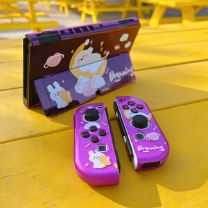 May include: A purple Nintendo Switch console with a custom design featuring a cartoon bunny and the word "Dreaming". The console is open and the two purple Joy-Con controllers are detached.