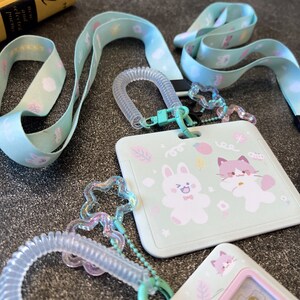 Op de afbeelding: Een licht muntgroene ID-badgehouder met een cartoonillustratie van een konijn en een kat, bevestigd aan een bijpassend stoffen koord. De set bevat een doorzichtige spiraalvormige polsband en iriserende stervormige bedels.
