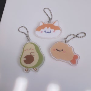 Op de afbeelding: Drie schattige sleutelhangers met cartoonfiguren: een groene avocado, een bruine en oranje kat en een gele gebakken garnaal. Elke sleutelhanger heeft een glimlachend gezicht en is bevestigd aan een zilveren ketting.