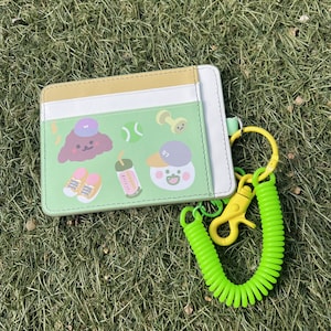 May include: A green card holder with a cartoon design featuring a dog, a drink, a tennis ball, and a dumbbell. The card holder has a yellow and white stripe and comes with a green coiled keychain.
