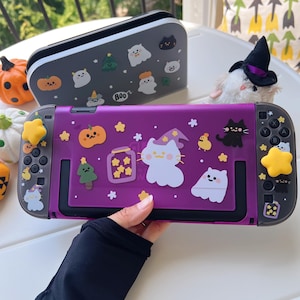 May include: A purple Nintendo Switch console with Halloween-themed stickers, including pumpkins, ghosts, and stars. A matching gray carrying case is in the background. The console is held by a person.