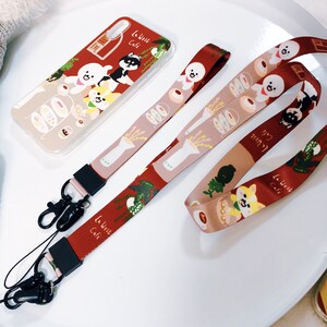 Może przedstawiać: Czerwono-biało-brązowe etui na telefon z kresk&oacute;wkowym wzorem psa i kota. Na etui widnieje napis "La West Cafe". Etui jest przymocowane do czerwono-biało-brązowego paska do telefonu z podobnym kresk&oacute;wkowym wzorem. Pasek ma dwa czarne metalowe klipsy.
