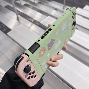 May include: A green and pink Nintendo Switch case with a cute cartoon design featuring cats, bunnies, and other animals. The case has a  "Catch Fun" slogan on it.