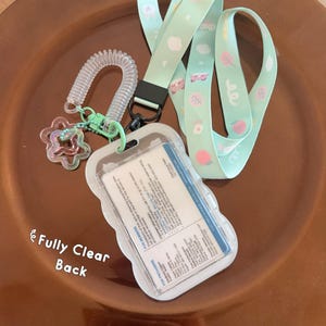 Op de afbeelding: Een licht mintgroen koord met een patroon van kleine witte wolkjes, roze blaadjes en kattengezichtjes. Het bevat een doorzichtige plastic badgehouder met een witte kaart erin, een spiraalvormige polsband en een stervormige bedel.