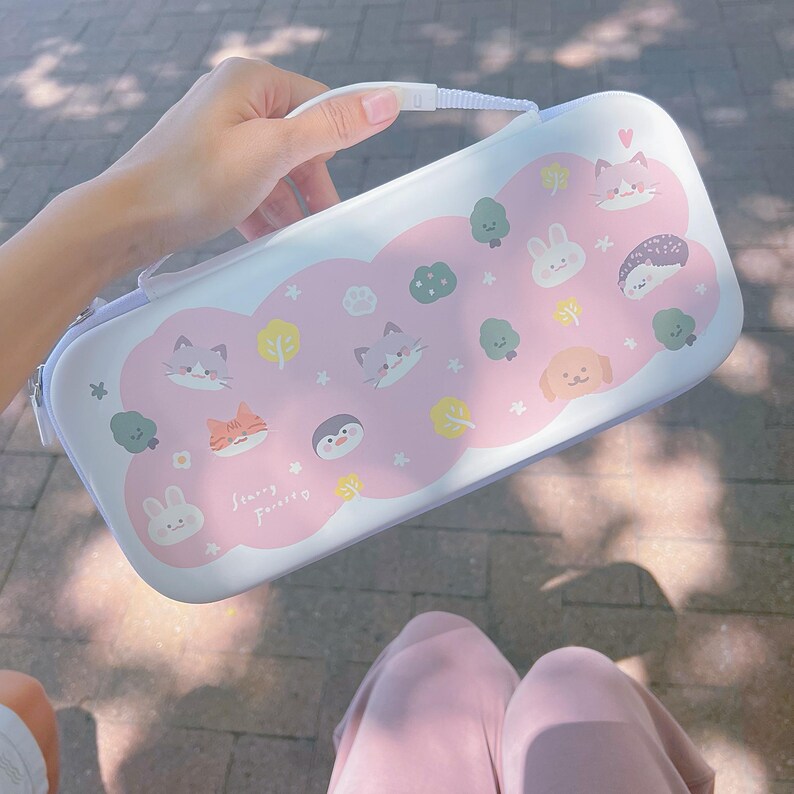 May include: A white and pink carrying case with a handle, featuring cartoon illustrations of animals and trees. The case has a light blue trim and the text "Sunny Forest".