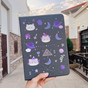 May include: A black galaxy-themed notebook cover with a purple and white illustration of a cat wearing a witch hat. The cover also features a purple triangle with the text "The Starry Forest Divination".