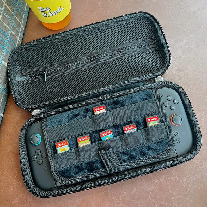 May include: Black zippered carrying case for a handheld gaming console, open to reveal the console and game cartridges. The case has a mesh top and a soft interior with slots for game storage. The console has black controls and a red button.
