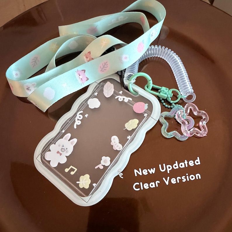 Op de afbeelding: Een licht mintgroen koord bevestigd aan een doorzichtige kaarthouder met golvende randen, versierd met kleine pastelkleurige stickers van een konijn, blaadjes en bloemen. Een transparant spiraalkoord verbindt twee stervormige bedels. Tekst: New Updated Clear Version.