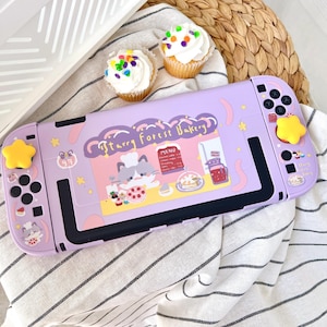 May include: A lavender Switch console case with a "Starry Forest Bakery" design, featuring a cat chef and menu illustrations. The case has yellow star-shaped buttons and is placed on a striped fabric with two cupcakes.