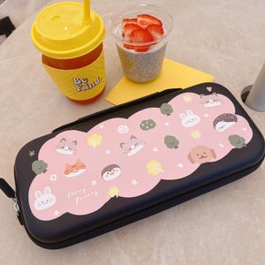 May include: Black pencil case with a pink cloud design featuring cartoon animals and the text "Starry Forest". A yellow drink with a straw and a cup of chia seed pudding with strawberries are also visible.