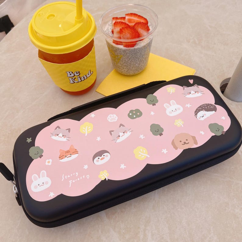 May include: Black pencil case with a pink cloud design featuring cartoon animals and the text "Starry Forest". A yellow drink with a straw and a cup of chia seed pudding with strawberries are also visible.