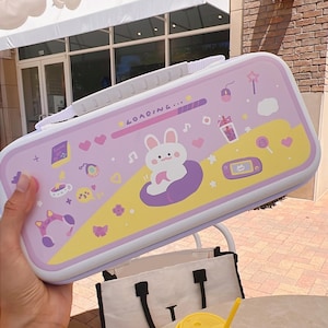 May include: A white ceramic mug with a black handle and the text "But First, Coffee" in a bold, black font. The mug is set against a blurred background. The mug appears to be approximately 10 cm tall.