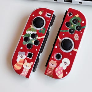 May include: Red and white Nintendo Switch Joy-Con controllers with a Christmas theme. The controllers feature a Christmas tree, gingerbread man, and animal illustrations. The buttons and joysticks are black.