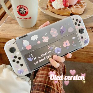 K&ouml;nnte beinhalten: Eine graue Nintendo Switch OLED-H&uuml;lle mit Blumen- und Vogelaufklebern. Auf der H&uuml;lle steht der Text "A Story of the Blue Sky". Eine Person h&auml;lt die H&uuml;lle. Im Hintergrund befinden sich eine Kaffeetasse und Waffeln.
