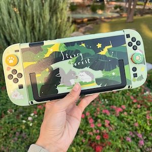 以下が含まれることがあります： リスとハリネズミが描かれた「Starry Forest」デザインのミントグリーンのNintendo Switchケース。ケースには黒い画面の縁とカラフルなボタンが付いています。左側には足跡とウサギのデザインがあります。