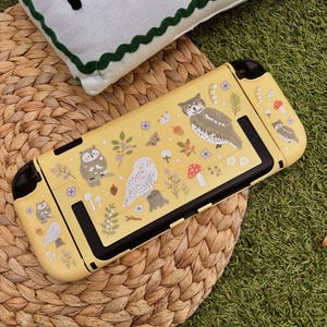 Pode incluir: Uma capa protetora amarela para console de jogos portátil com um padrão de corujas, cogumelos vermelhos, pequenas flores e folhagem verde. A capa tem detalhes pretos e aberturas para botões e portas, com um acabamento fosco e suave.