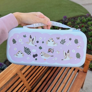 以下が含まれることがあります： 白のハンドルとジッパー開閉部が付いた、水色のキャリングケース。ケースには、白、茶色、緑の色合いの鳥、葉、ベリーの繰り返し模様が、薄紫色の背景に描かれています。