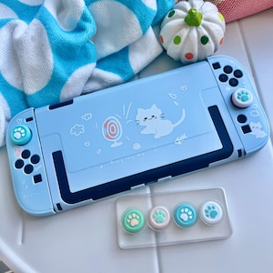 May include: A light blue Nintendo Switch console with a cat and fan design. The console has matching paw print thumb grips and additional thumb grips in a clear case. A blue and white towel and a decorative pumpkin are in the background.