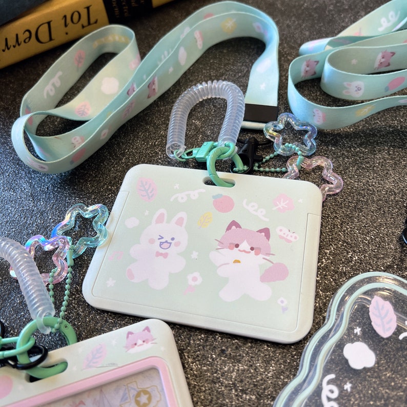 Op de afbeelding: Een licht mintgroene plastic badgehouder met een cartoonillustratie van een konijn en een kat, bevestigd aan een bijpassend mintgroen koord met een transparant spiraalsnoer en iriserende stervormige bedels voor veilige identificatie.