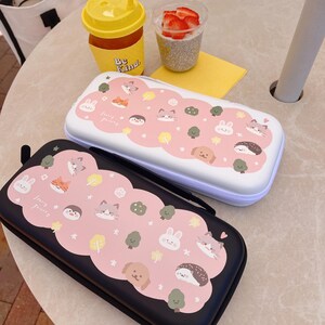 May include: Two rectangular cases, one black and one white, with a pink cloud design. The cloud design features cartoon animals, trees, and stars. The cases have a zipper closure and a handle. The cases are likely for storing small items.