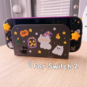 Pode incluir: Uma consola Nintendo Switch preta e roxa com um suporte decorativo. O suporte apresenta um design temático de Halloween com gatos de desenho animado, abóboras, estrelas e o texto "For Switch 2". A consola tem decorações amarelas em forma de estrela.