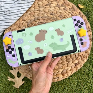 Puede incluir: Una funda verde menta para Nintendo Switch con capibaras de dibujos animados, tréboles y un diseño de cocodrilo. La funda tiene detalles en morado y cubiertas de joystick en forma de estrella amarilla. La Switch está sobre una superficie de mimbre tejido.