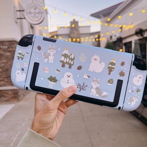 以下が含まれることがあります： 冬をテーマにしたステッカーで飾られた、水色のNintendo Switchケース。漫画の動物、木、雪だるまなどが含まれています。ケースには黒いアクセントがあります。