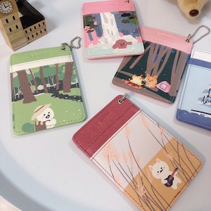 May include: Five rectangular card holders with metal ball chains, each featuring distinct illustrations of cartoon characters in nature settings, including a forest, a waterfall, a campfire, and a field of tall grass with a character playing a guitar.