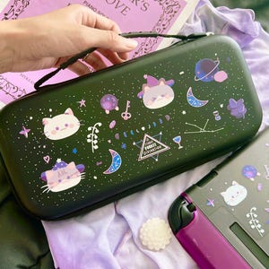 以下が含まれることがあります： 黒い持ち運び用ケース。猫と宇宙をテーマにしたステッカーで装飾されています。ケースにはジッパー開閉と黒いハンドルが付いています。ステッカーには、猫、惑星、月、星、そして「STARRY FOREST DIVINATION」の文字が含まれています。