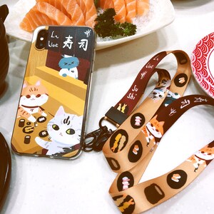 Może przedstawiać: Brązowe i złote etui na telefon z kresk&oacute;wkową ilustracją kot&oacute;w jedzących sushi. Etui zawiera napis "La Wei" i "寿司" w chińskich znakach. Etui jest połączone z brązowym i złotym sznurkiem w stylu sushi.